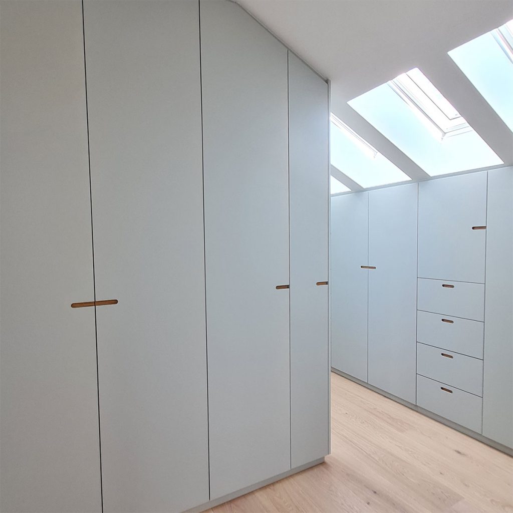 Blick in eine Ankleide im Dachgeschoss- links und rechts Einbauschränke mit grau lackierten Fronten und Muldengriffen, die mit Holz hinterlegt sind. Der Fußboden ist mit hellem Eichenparkett belegt.