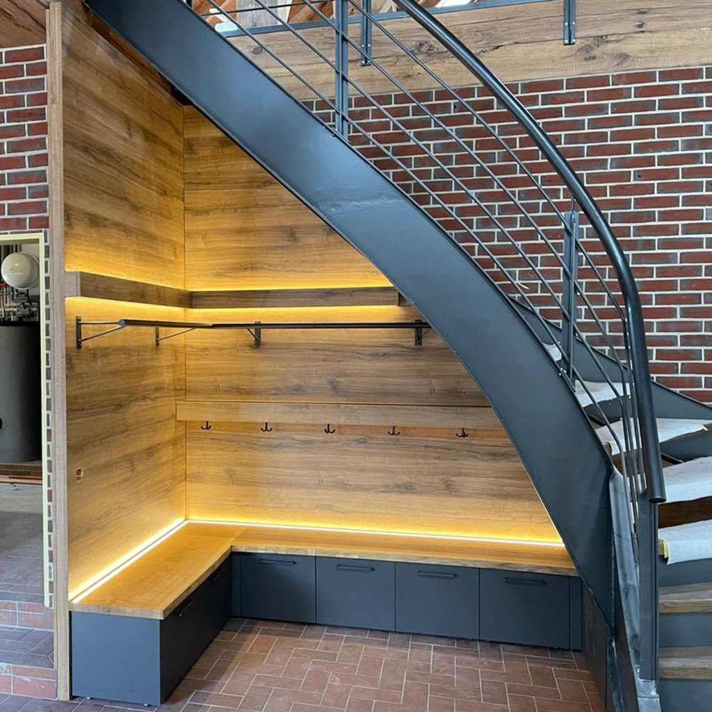 Blick auf eineGarderobe unter einer Treppe– unten eine Sitzbank aus einer massiven Eichenholzplatte, darunter Rollschränke mit dunkelgrauen Fronten. Die Wände in der Garderobe sind mit Möbelplatte in rustikal Holzdekor verkleidet. Garderobenstange und Garderobenhaken aus Schwarz lackiertem Stahl. Der Fußboden ist mit roten Fliesen belegt.