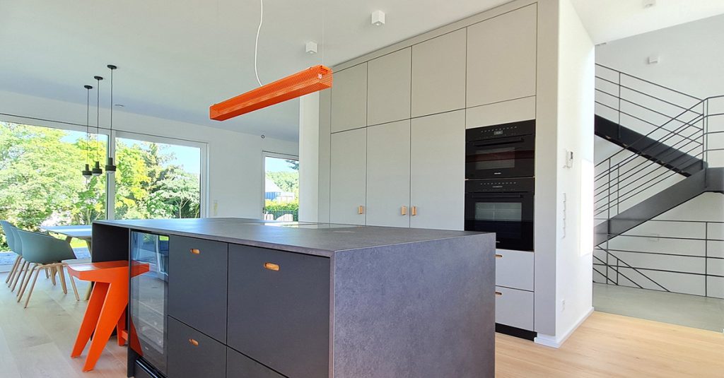 Blick in eine offene Küche – vorne steht zentral ein schwarzer Küchenblock mit Arbeitsplatte und Seiten aus Quarzstein, die Küchenfronten ausgeführt in schwarzem Linoleum mit Muldengriffen, die mit Holz hinterlegt sind. Dahinter steht eine Hochschrankzeile wie ein Raumteiler. Die Hochschrank Zeile hat hellgraue Möbelfronten. Über dem Küchenblock hängt eine orangene Pendelleuchte. Links neben der Hochschrank Zeile schaut man in das offene Wohnzimmer und in den dahinterliegenden Garten. Rechts daneben blickt man in das offene Treppenhaus.