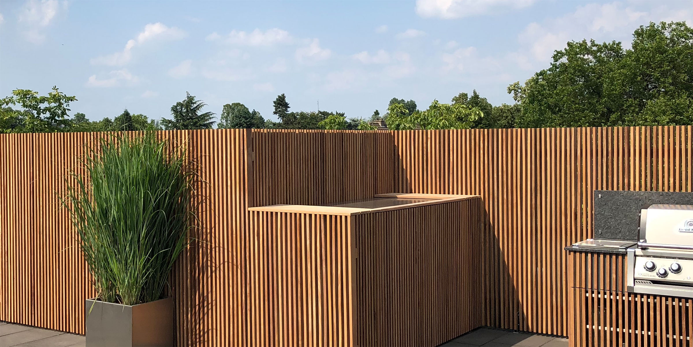 Sichtschutzwände und integrierte Outdoorschränke in Lamellenoptik gefertigt aus Terrassenholz, rechts anschließend eine Outdoorküche mit Gasgrill in Edelstahl . Links vor der Lamellenwand steht eine grüne Pflanze in einem Kübel aus Edelstahl.