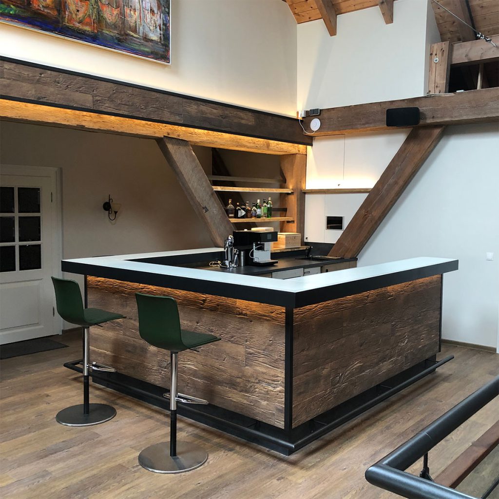 Hausbar eingebaut in einer alten Scheune, Bar in rustikalem Still mit Thekenfronten in Altholz und Thekentop aus beleuchtetem Glas, Einfassung des Thekentops und Fußstütze aus schwarz lackiertem Stahl, davor stehend 2 Barhocker