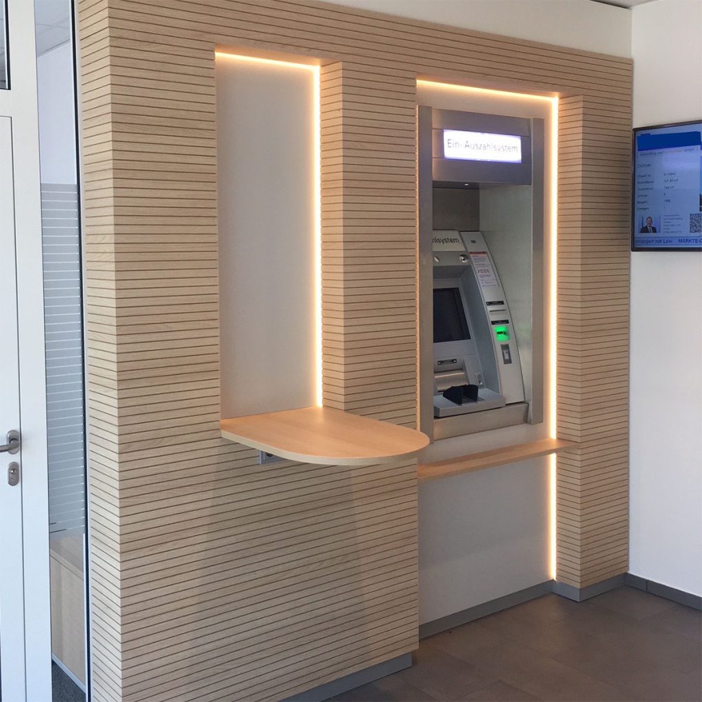 Blick in die Service. Filiale einer Volksbank – zu sehen, ist eine Wandverkleidung in Lamellenoptik gefertigt aus hellem Holz. Rechts ein integrierter Geldautomat links ein kleiner Besprechung Tisch, der an der Wand befestigt ist.