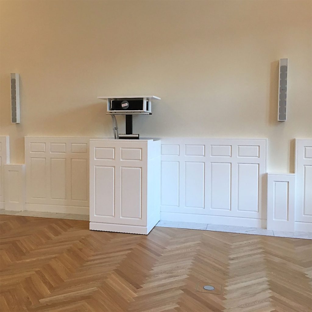 Aufnahme in einem historischen Konferenzsaal – zu sehen, ist eine Wandverkleidung mit Rahmen und Füllungen in weiß. Passend zur Wandverkleidung ein Medienschrank, aus dem ein Beamer herausgefahren ist. Über der Wandverkleidung eine beige gestrichene Wand mit zwei Lautsprechern. Fußboden als Fischgrätparkett in Eiche mit umlaufendem Fries in Marmor.