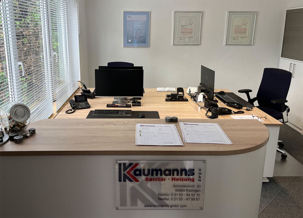 Blick in das helle Büro eines Handwerksbetriebs – vorne ein kleiner Tresen mit weißer Front und Oberplatt in Holz. Dahinter eine Schreibtischinsel mit drei Arbeitsplätzen in der gleichen Farbkombination. An der Wand hängen die Meister Briefe des Unternehmens.