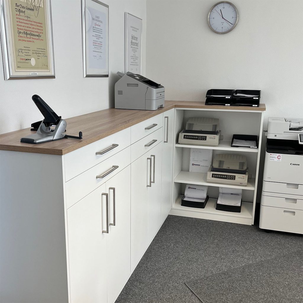 Büro in einem Handwerksbetrieb – links ein Sideboard als Aktenschrank rechts ein offenes Regal für Drucker. Das Sideboard ist gefertigt aus weißer Möbelplatte mit einer Deckelplatte aus Holz.