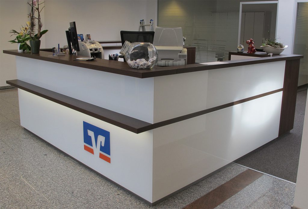 Empfangtresen in einer Volksbank in U-Form- Thekenfronten aus weißem Glas mit einer Taschenablage und einem Thekenbrett in dunkelbraunen Holz. Unter der Taschenablage befindet sich eine indirekte Beleuchtung und das Logo der Volksbank.