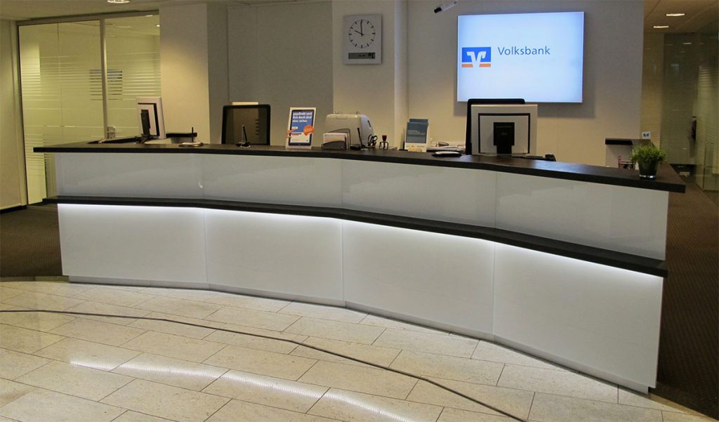 Empfangtresen in einer Volksbank – breiter Tresen mit Konkaver Front– Thekenfront aus weißem Glas. Thekenbrett und Taschenablage aus dunklen Holz. Unter der Taschenablage ist eine indirekte Beleuchtung. Hinter dem Tresen befinden sich zwei Arbeitsplätze.