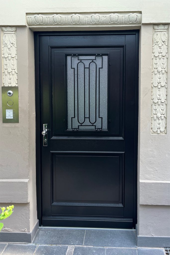 Schwarze Haustüre aus Holz eingebaut in eine Gründerzeitfassade. Haustüre mit klassischen Füllungen und Profilen oben mit Lichtausschnitt und schwarzem Stahlgitter. Verglasung, Ornament,glas Eisblume. Sicherheitsbeschlag. Bauhaus Edelstahl poliert.