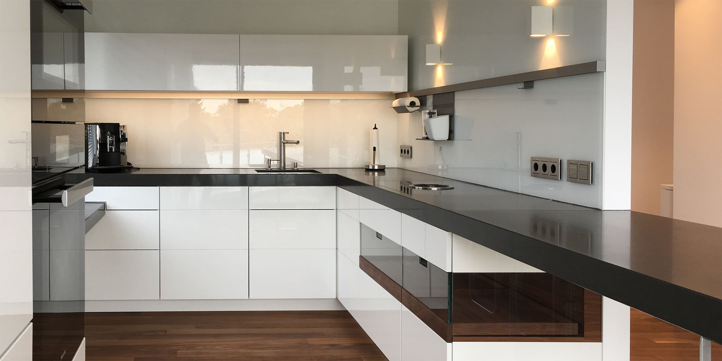 Blick in eine Einbauküche. Unterschränke. Hochglanz weiß lackiert, teilweise mit Fronten aus Glas. Arbeitsplatte aus schwarzem Quarzstein mit eingebautem Bora Kochfeld. Küchenspiegel aus weißem Glas und einem Hängeschrank Weiß glänzend lackiert.