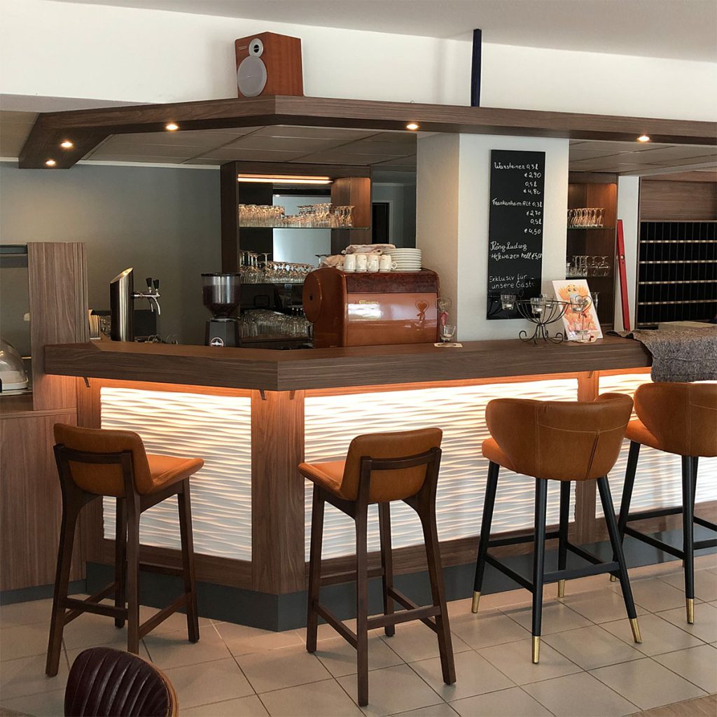 Blick in eine Hotelbar – das Bild zeigt eine Theke mit Fronten aus Reliefplatte eingesetzt in einer Rahmenkonstruktion. Rahmenkonstruktion und Thekenbrett aus dunklem Holz. Unter dem Thekenbrett eine indirekte Beleuchtung. Hinter der Theke, eine Zapfanlage und eine große Kaffeemaschine. Vor der Theke drei Barhocker.