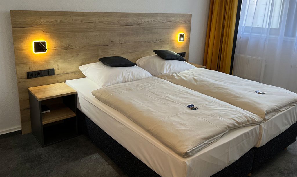 Blick in ein Hotelzimmer – ein Doppelbett, bestehend aus zwei Boxspringbetten, dahinter ein großes Kopfteil aus Möbelplatte in hellem, rustikalen Holzdekor mit zwei Lampen. Links ein Nachtschrank mit Oberplatte in Holz und Seiten in dunkelgrau.