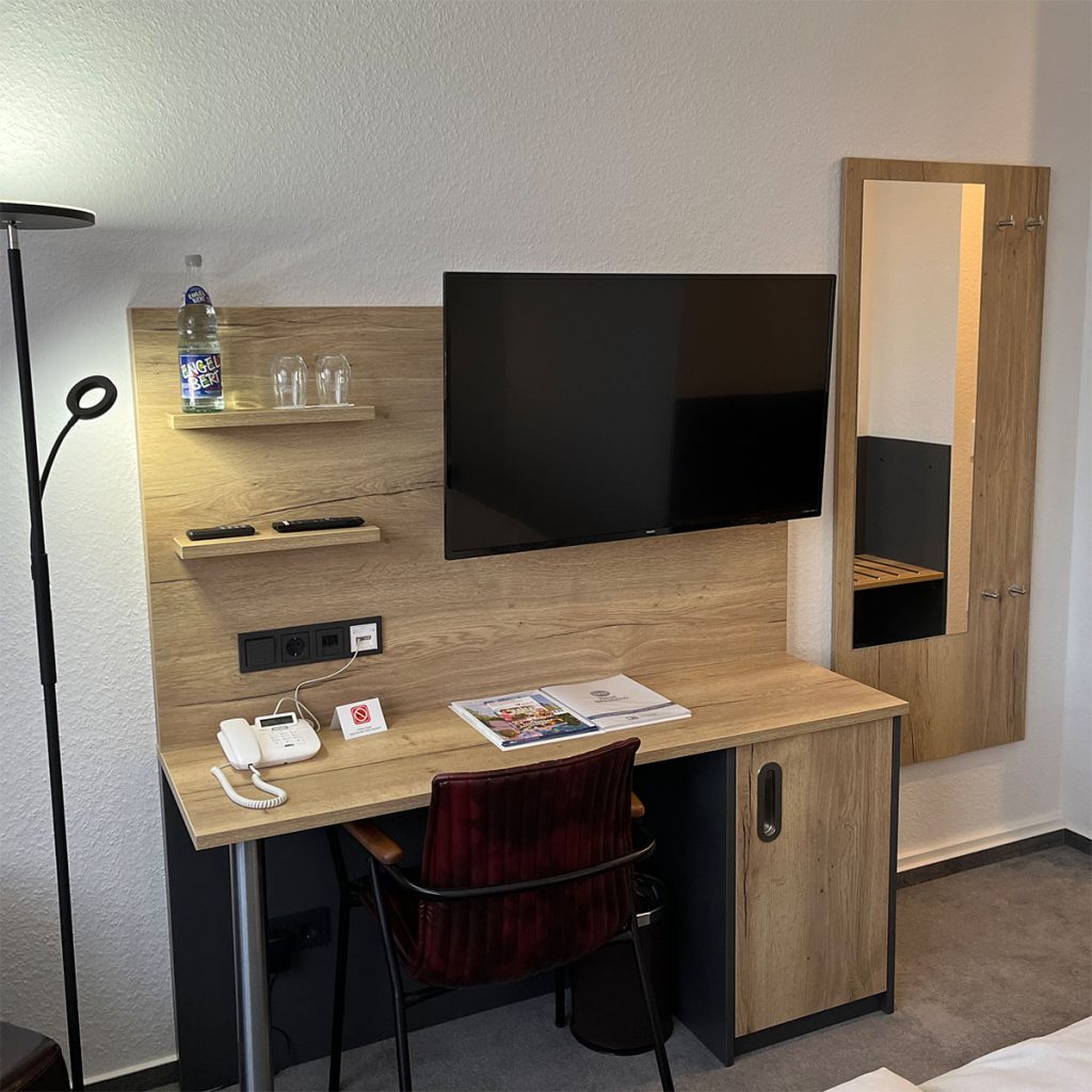 Detailaufnahme in einem Hotelzimmer – das Bild zeigt ein Kombinationsmöbel bestehend aus einem Schreibtisch mit einer TV Wand. Der Schreibtisch hat einen Unterschrank mit Türe. An der TV Wand sind zwei Ablagebretter und ein TV Gerät montiert. Rechts neben dem Möbel hängt ein Garderobenboard mit Spiegel und vier Garderobenhaken. Alle Möbel sind aus Möbelplatte in hellem Holzdekor gefertigt.