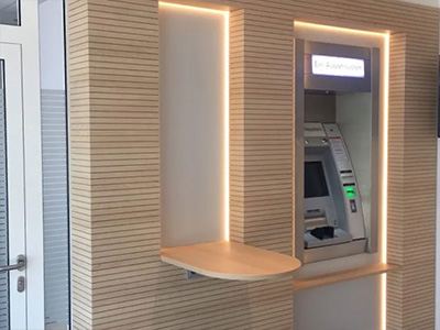 Blick in den Service Point einer Bank Filiale – zu sehen ist eine Wand, die in waagerechter Lamellenoptik verkleidet ist. Die Verkleidung ist aus hellem Ahornholz gefertigt.Rechts steht ein Geldautomat links ragt eine abgerundete Tischplatte aus der Wandverkleidung.
