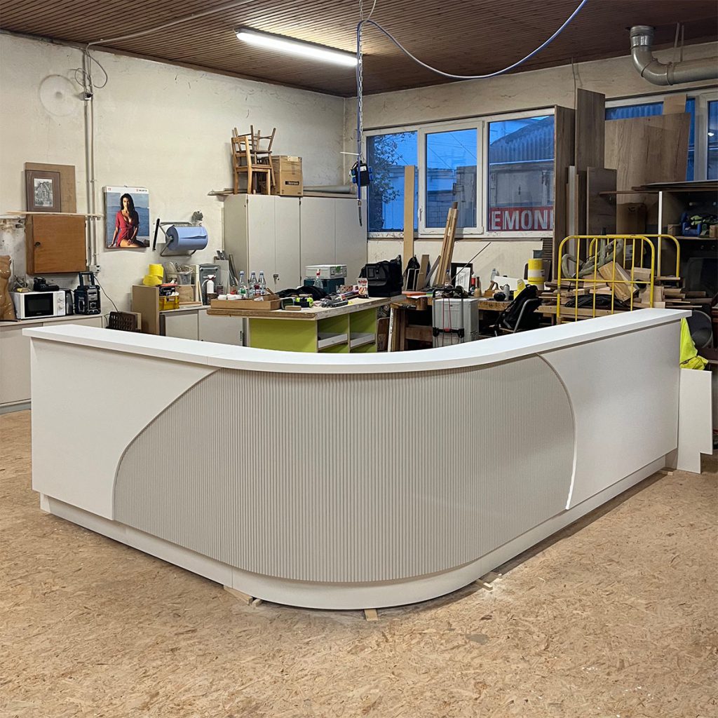 . Einblick in die Werkstatt. Ein Praxistresen in L Form..Die Ecke ist als viertel Kreis Rundung ausgeführt. Die Thekenfront in diesem Bereich aus grau lackierten Lamellen. Rechts und links daneben glatte Thekenfronten in weiß. Thekenbrett in weiß.