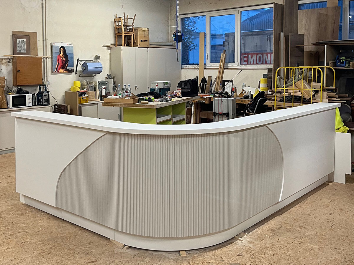 Ein maßgefertigter Empfangstresen in L-Form, Ecke vorne als Viertelkreisrundung in Lamellenoptik, gefertigt mit modernen Design-Elementen in weiß und grau in hochwertiger Verarbeitung. Tresen steht noch in der Werkstatt