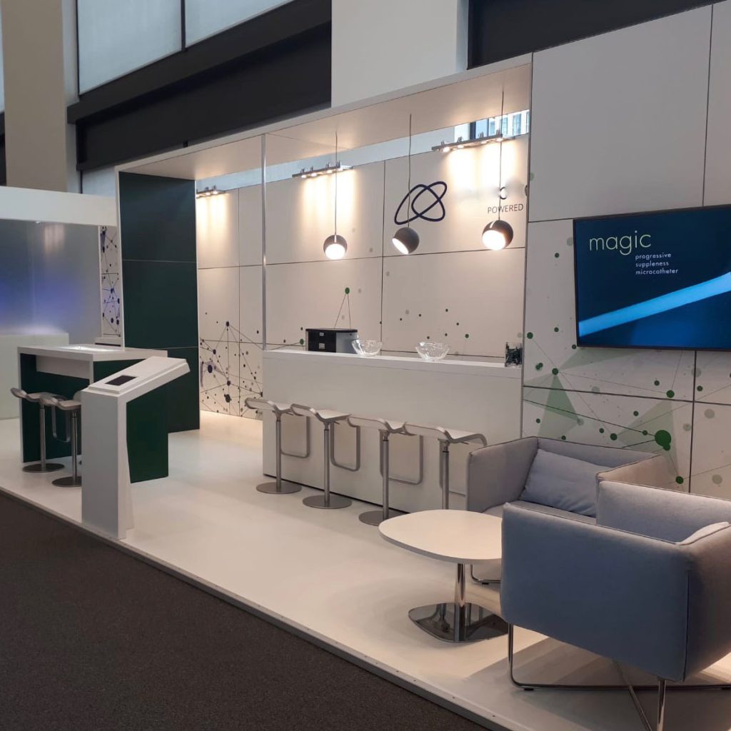 Blick auf einen Messestand in weiß. In der Mitte steht ein schlichter Tresen. Mit vier Barhocker. Darüber hängen drei Lampen. Rechts vor dem Tresen steht eine kleine Sitzgruppe mit zwei Sesseln und einem Bistrotisch. Vorne an der Stand Cante steht ein Infoboard mit einem Tablet.