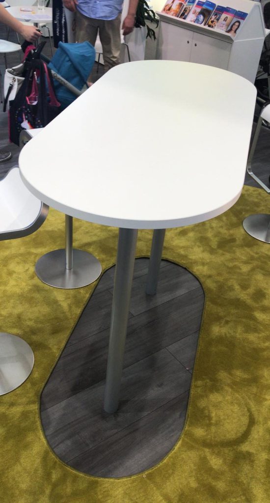 Stehtisch auf einem Messestand. Eine längliche Tischplatte mit zwei Viertel-Kreisrundungen gefertigt aus weißer Möbelplatte. Zwei Tisch Beine aus Stahlrohr Silber lackiert. Ein Tischfuß in der gleichen Form der Tischplatte aus Möbelplatte in Holzdekor.