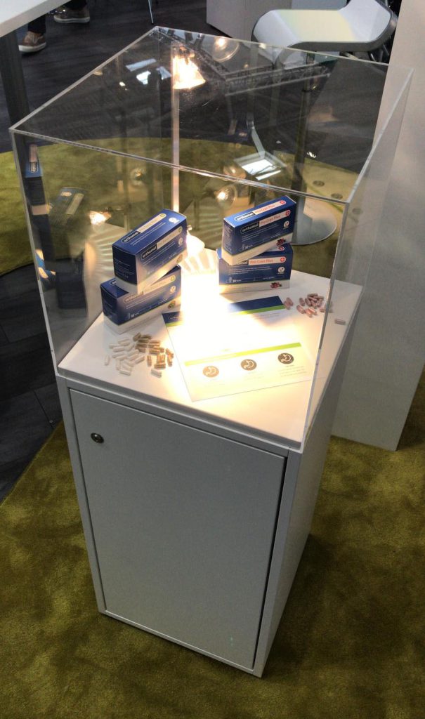 Blick auf eine Messevitrine – ein weißer Unterschrank mit abschließbare Türe, darauf eine Haube aus Plexiglas. In der Vitrine liegen Exponate, die beleuchtet werden. Der Schrank steht auf einem grünen Teppich.