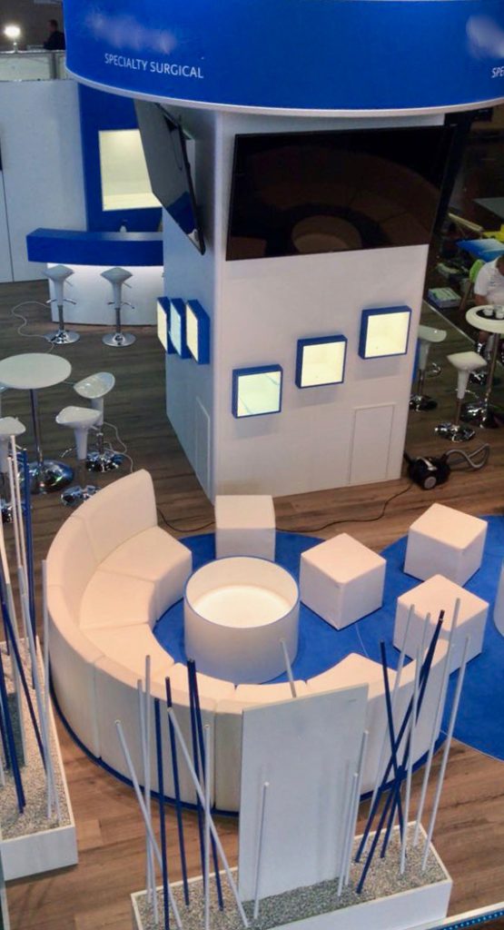 Blick von oben auf einen Messestand. Im Zentrum steht eine halbe Runde Sitzgruppe um einen runden Tisch. Im Hintergrund ein zentraler Turm von zwei Seiten zu sehen mit jeweils drei beleuchteten Vitrinen in blau.