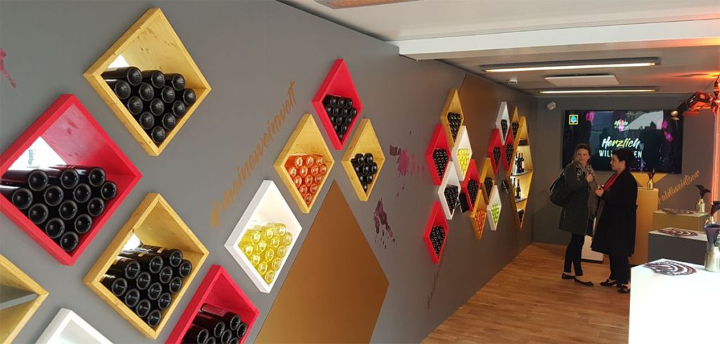 Blick in einen Pop-up Store. Zu sehen, ist eine Produktwand, in die mehrere quadratische Korpusse eingebaut sind. Die Korpusse haben verschiedene Farben – hier rot, gelb und weiß. Die Wand ist grau mit goldener Schrift. In den Korpussen liegen Weinflaschen wie in einem Weinkeller.