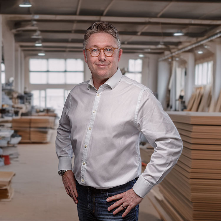Daniell Cremerius Schreinermeister und Inhaber der Kröll und Cremerius Einrichtungs GmbH. Er steht in der Werkstatt und trägt Jeans sowie ein weißes Hemd.