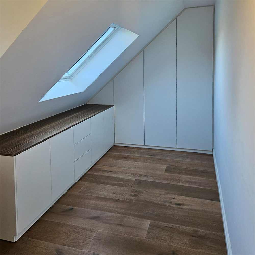 Einbauschrank unter Dachschräge mit 3 Türen. Links anschließend ein Sideboard unter einem Dachfenster. Sideboard mit Oberplatte in dunkelbraunem Eichenholz passend zum Parkettboden. Möbelfronten ohne Griffe, weiß lackiert