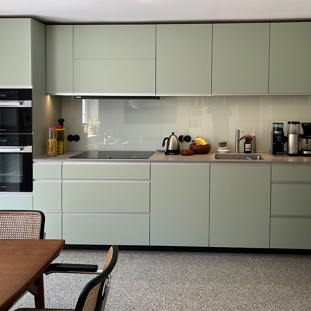 Pastellgrüne Küchenzeile links mit Gerätehochschrank rechts mit Unterschränken und Hängeschränken. Fonten in Pistaziengrün mit Profilgriffen in Aluminium. Arbeitsplatte aus Quarzstein in Betonoptik, Küchenspiegel aus hinterlackiertem Glas