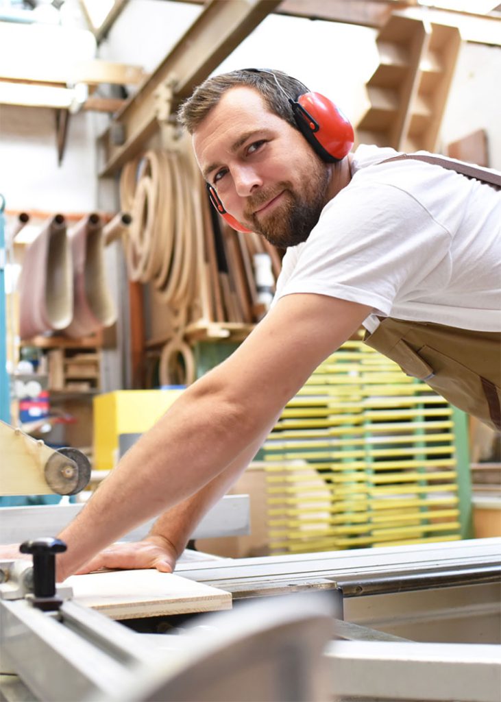 Mitarbeiter mit Gehörschutz lächelt in die Kamera, während er an der Formatkreissäge ein Stück Holz zusägt.
