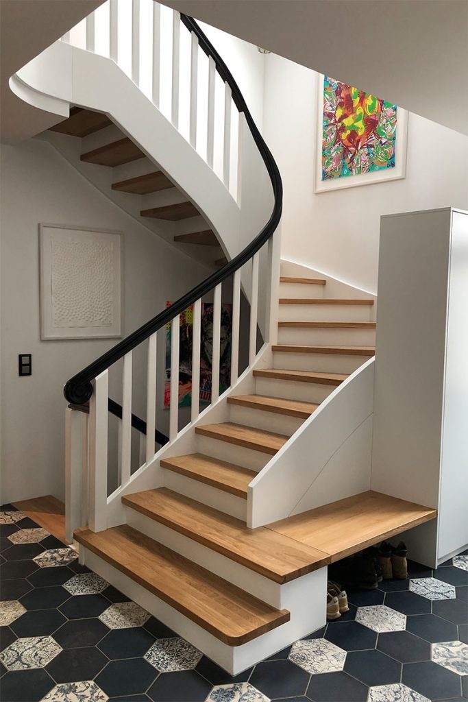 Blick in ein Treppenhausauf eine Halb gewendete Treppenanlage. Klassische Wangentreppe mit eingestemmten Stufen aus Eiche. Wangen und Treppengeländer weiß lackiert. Durchlaufender Handlauf in Omegaform in schwarz. Rechts angrenzend eine Sitzbank in Eiche und ein Einbauschrank in weiß lackiert.