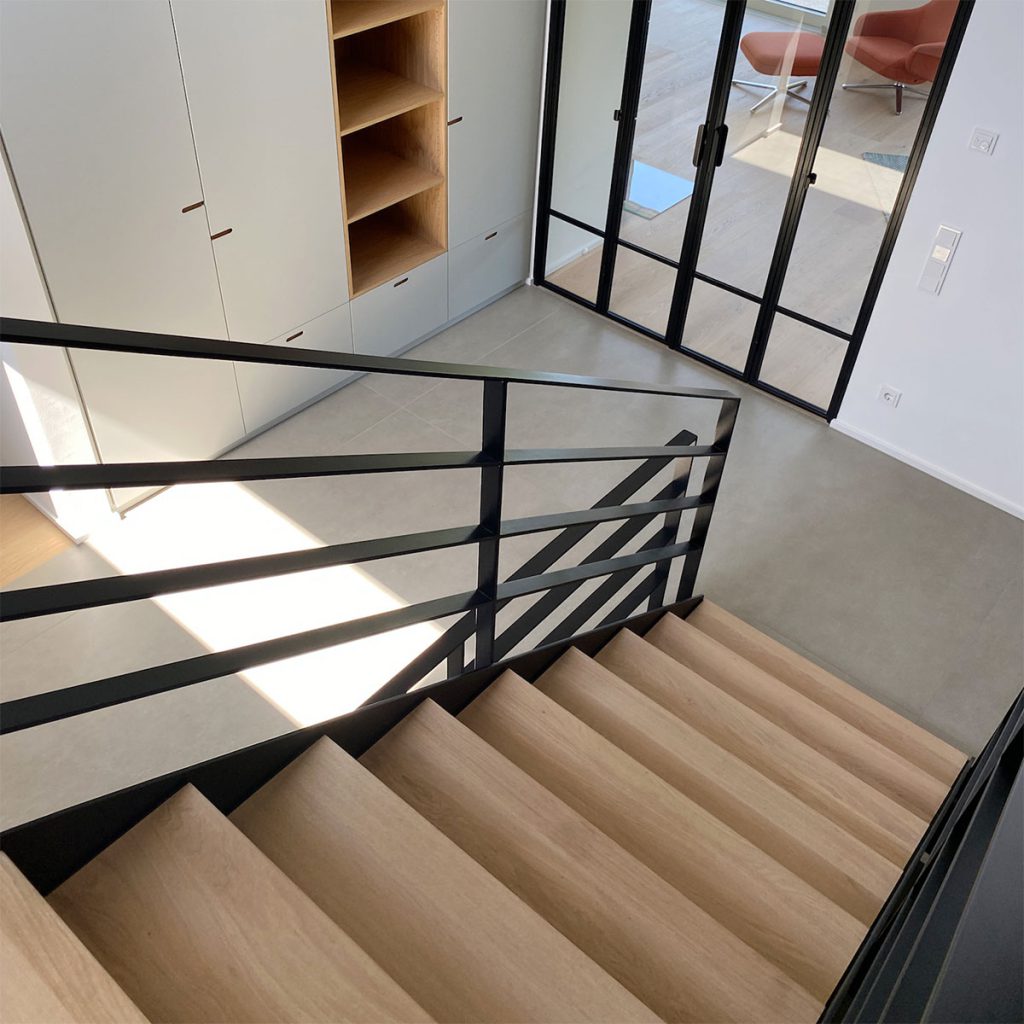 Detail einer Treppenanlage. Stufen in Massivholz. Eiche weiß geölt. Treppengeländer im Industriestil aus Schwarz lackiertem Stahl. Im Hintergrund ein Einbauschrank und eine Glastüre im Loftstil.
