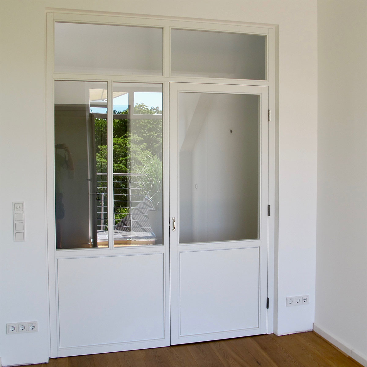 WeißeTüranlage im schlichten geradlinigen Stil, eingebaut in einer Wohnung mit Parkettboden und weißen Wänden. Links ein festes Element. Unten mit geschlossener Füllung oben mit Festverglasung und einem Mittelsteg. Oben ein Oberlicht mit zwei Lichtfeldern. Rechts Türflügel unten mit geschlossener Füllung oben mit Verglasung. Verglasung als Klarglas ausgeführt.